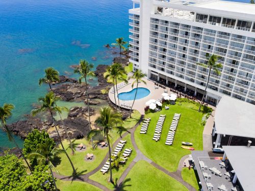Aerial view of a beachfront hotel with a pool, palm trees, and lounge chairs by the ocean.