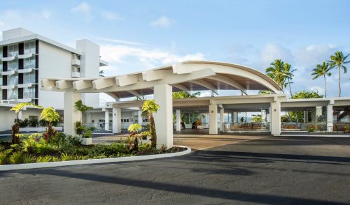 Modern hotel entrance with a curved roof, surrounded by palm trees and landscaped gardens under a clear blue sky.