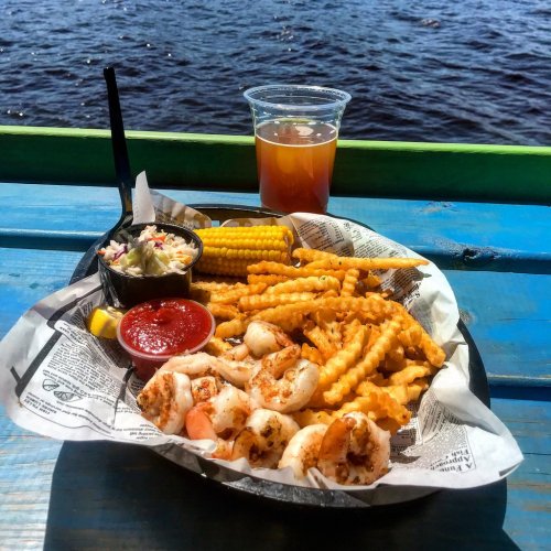 A basket of shrimp, fries, coleslaw, and dipping sauces, with a drink, set against a water backdrop.