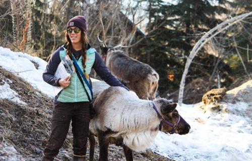 A person in winter attire stands beside a reindeer in a snowy landscape, with trees in the background.