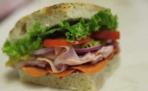 A close-up of a sandwich with layers of lettuce, tomato, onion, ham, pepperoni, and pickles on soft bread.