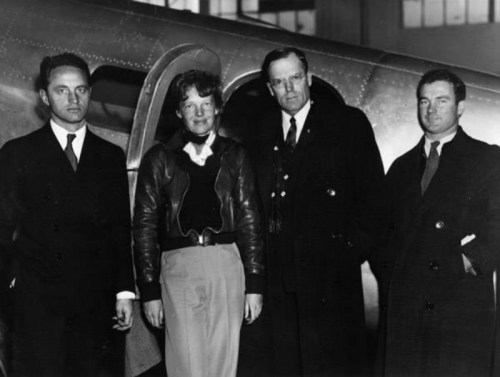 Four people in formal attire stand in front of an airplane, with a vintage aviation backdrop.
