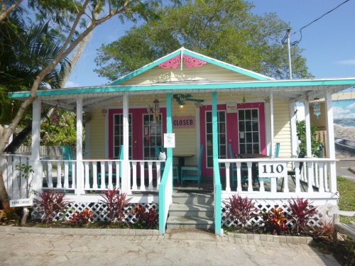 A colorful, closed cottage with pink doors and a porch, surrounded by greenery and tropical plants.