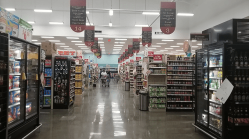 A grocery store aisle with shelves of products, coolers on the sides, and signs indicating different sections.