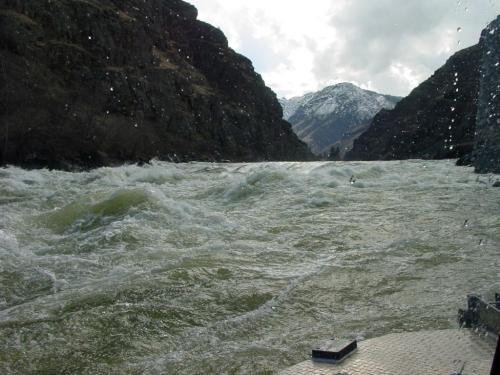 Raging river flows between steep, rocky cliffs under a cloudy sky, with snow-capped mountains in the background.