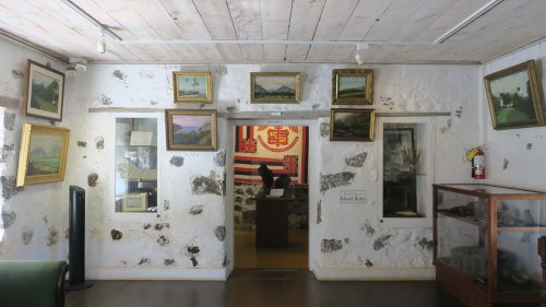 Interior of a museum room with framed paintings, a flag, and display cases against a stone wall.
