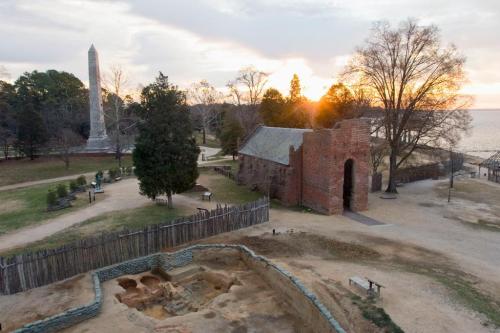 Aerial view of a historic site with ruins, a monument, trees, and a sunset over a body of water in the background.