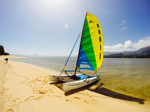 A colorful Hobie catamaran with a sail on a sandy beach, near calm waters and mountains in the background.