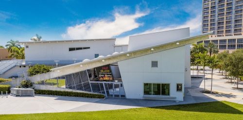 Modern building with a sloped roof, large windows, and a sign for "Hot Glass" surrounded by green grass and palm trees.