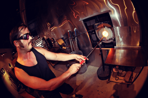 A glassblower in sunglasses shapes molten glass with a rod in a workshop, surrounded by tools and equipment.
