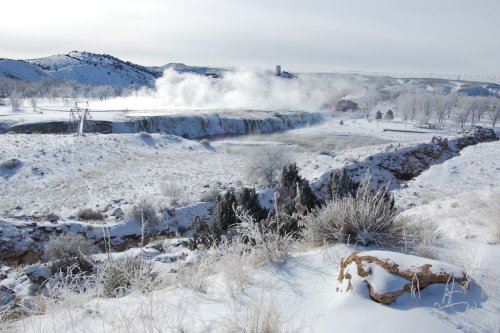 A snowy landscape featuring a waterfall with steam rising, surrounded by frosted trees and distant hills.