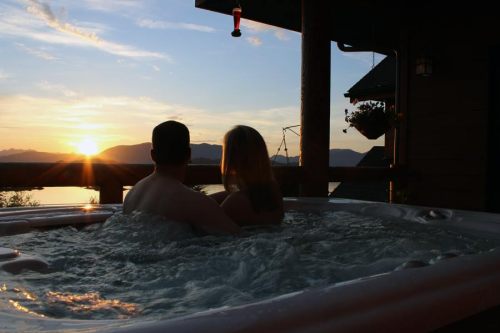 A couple relaxes in a hot tub, enjoying a sunset view over the water and mountains.
