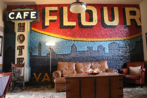 A cozy cafe interior featuring a vintage "Flour" mural, a brown leather couch, and a wooden coffee table.