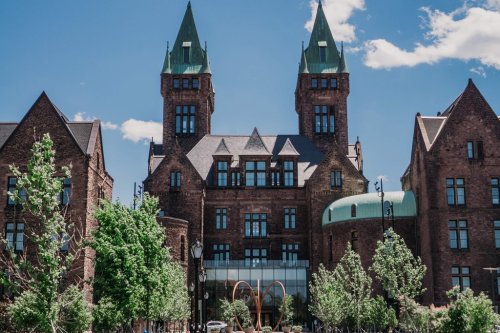 Historic brick building with green spires, surrounded by trees and a clear blue sky.