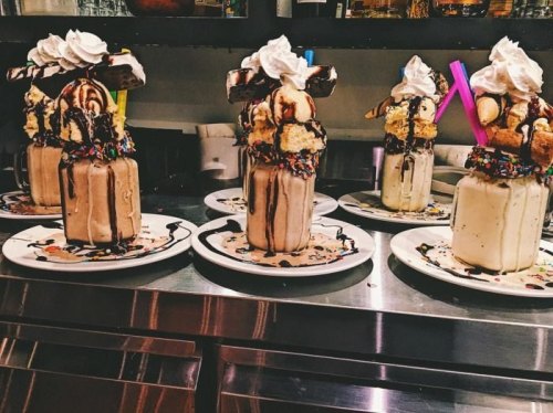 Four extravagant milkshakes topped with whipped cream, chocolate, sprinkles, and cake slices on white plates.