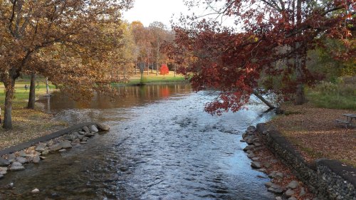 A serene river flows through a park with autumn foliage, featuring trees with vibrant red and orange leaves.