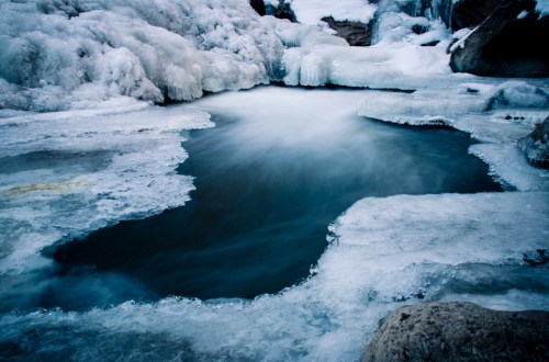 A serene, icy landscape featuring a partially frozen pool surrounded by snow-covered rocks and ice formations.