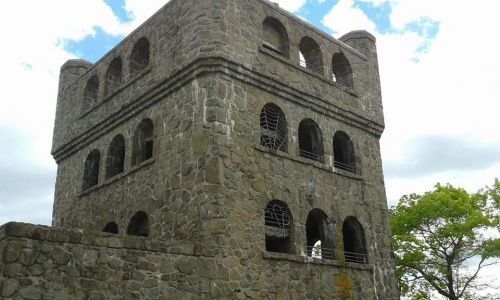 A stone tower with arched windows and a cloudy sky in the background, surrounded by greenery.