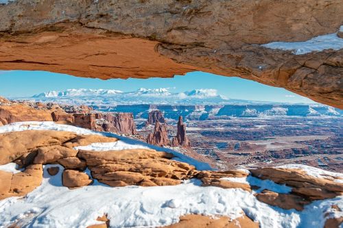 A snowy landscape framed by a rock arch, showcasing distant mountains and rugged terrain under a clear blue sky.