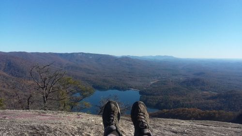 A pair of hiking shoes resting on a rocky ledge, overlooking a valley and lake surrounded by mountains under a clear blue sky.