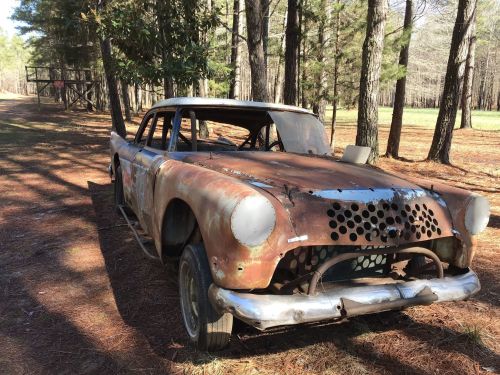 An old, rusty car sits among tall pine trees, showcasing peeling paint and a weathered exterior.