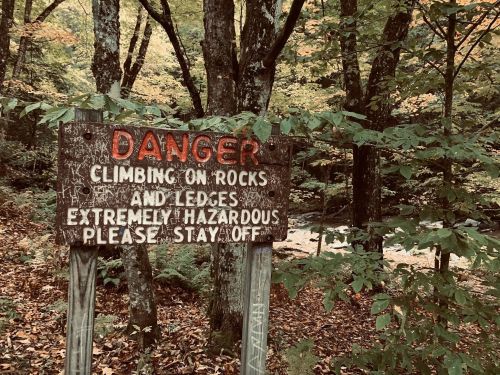 A weathered sign warns of hazards from climbing on rocks and ledges in a wooded area.