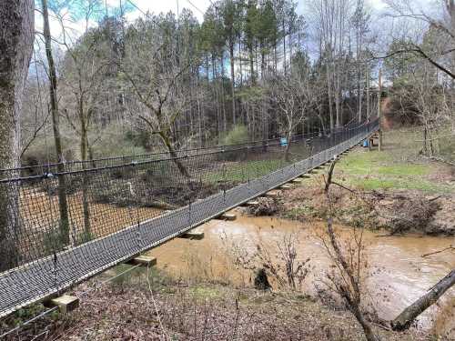 A suspension bridge spans a creek, surrounded by trees and a grassy area under a cloudy sky.