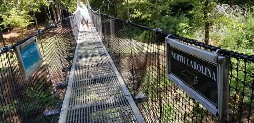 A person walks on a suspension bridge with signs indicating "North Carolina" on either side, surrounded by trees.