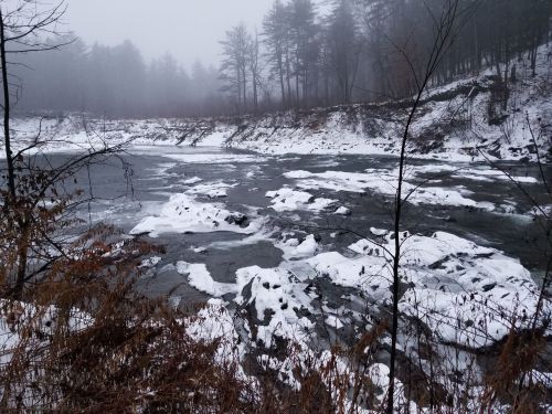 A foggy river scene with snow-covered rocks and trees lining the banks, creating a serene winter landscape.