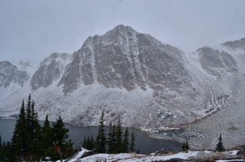 A snowy mountain landscape with a lake in the foreground and evergreen trees, under overcast skies.