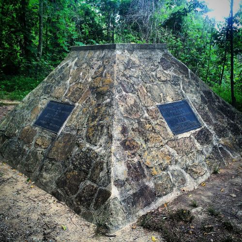 A stone monument with two plaques, surrounded by greenery in a wooded area.