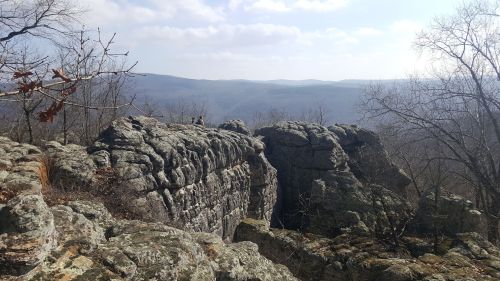 A rocky outcrop with trees, overlooking a valley and distant mountains under a partly cloudy sky.