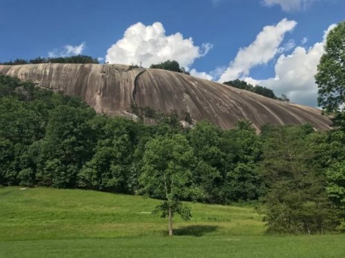 A large, smooth rock formation rises above a green landscape under a blue sky with fluffy white clouds.