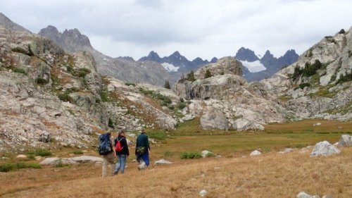 Two hikers walk through a rocky landscape with mountains in the background under a cloudy sky.