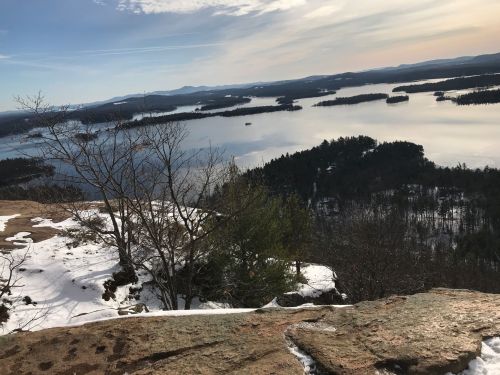A scenic view from a rocky overlook, featuring a lake surrounded by trees and distant mountains under a clear sky.
