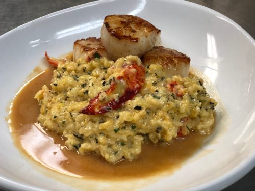 A plate featuring three seared scallops atop creamy risotto with lobster, drizzled with a rich sauce.