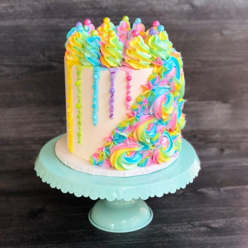 A colorful cake with swirled pastel frosting, topped with decorative peaks and drips, displayed on a mint green cake stand.