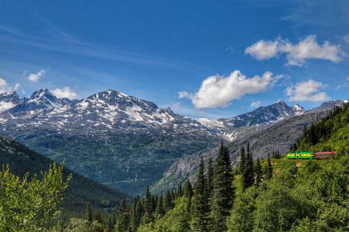 A colorful train travels through a lush valley surrounded by majestic snow-capped mountains and a clear blue sky.