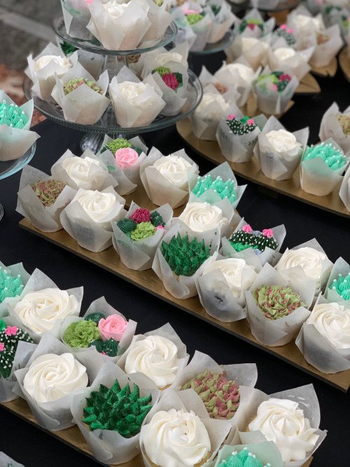 A display of beautifully decorated cupcakes in floral and cactus designs, arranged on wooden trays.