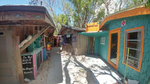 Colorful wooden buildings line a narrow walkway, surrounded by trees and signs, creating a vibrant outdoor space.