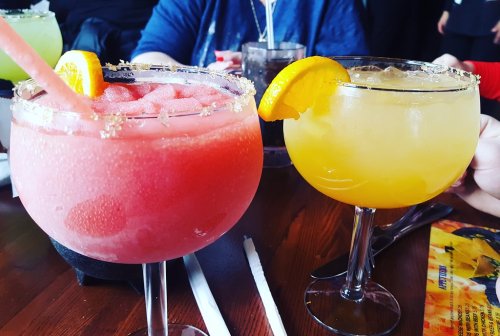 Two colorful cocktails on a wooden table: a pink frozen drink with a lemon slice and a yellow drink with an orange slice.