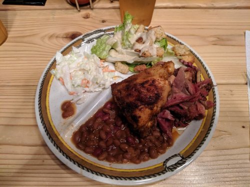 A plate of food featuring baked chicken, pulled pork, baked beans, coleslaw, and a side salad on a wooden table.