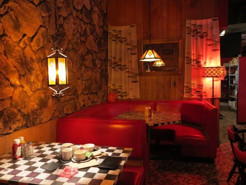 Cozy diner booth with red seating, checkered table, and warm lighting from lamps and a lantern against a wooden wall.