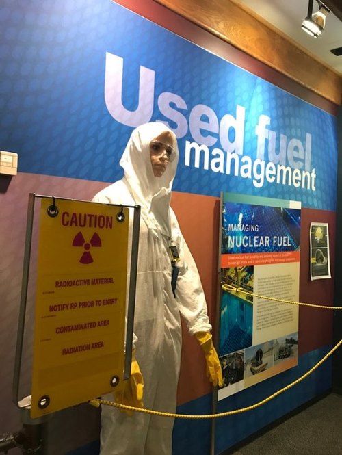 A person in a protective suit stands near a sign about used fuel management and radioactive material safety.