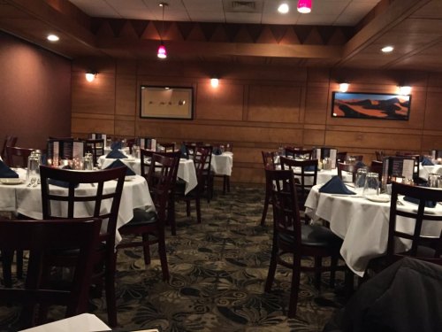 A dimly lit restaurant with empty tables set with white tablecloths and blue napkins, featuring wooden decor.