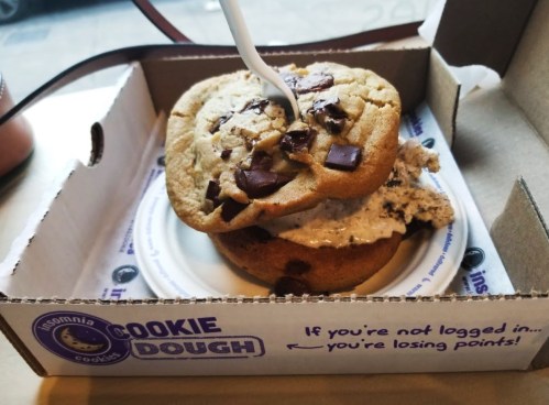 A cookie sandwich with chocolate chip cookies and cookie dough ice cream, served in a takeout box.