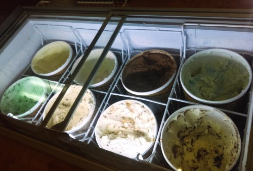 A display freezer with eight tubs of various ice cream flavors, including green mint, chocolate, and vanilla.