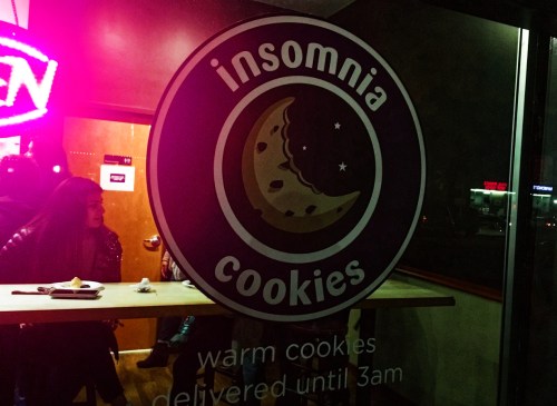 Logo of Insomnia Cookies on a window, featuring a cookie graphic and text about warm cookie delivery until 3 AM.