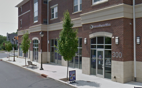 Exterior view of a brick building featuring Insomnia Cookies, with trees and a bench in front.