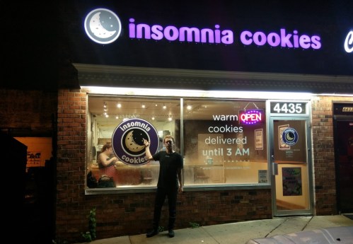 A person stands outside Insomnia Cookies, a brightly lit shop with a sign indicating it's open until 3 AM.